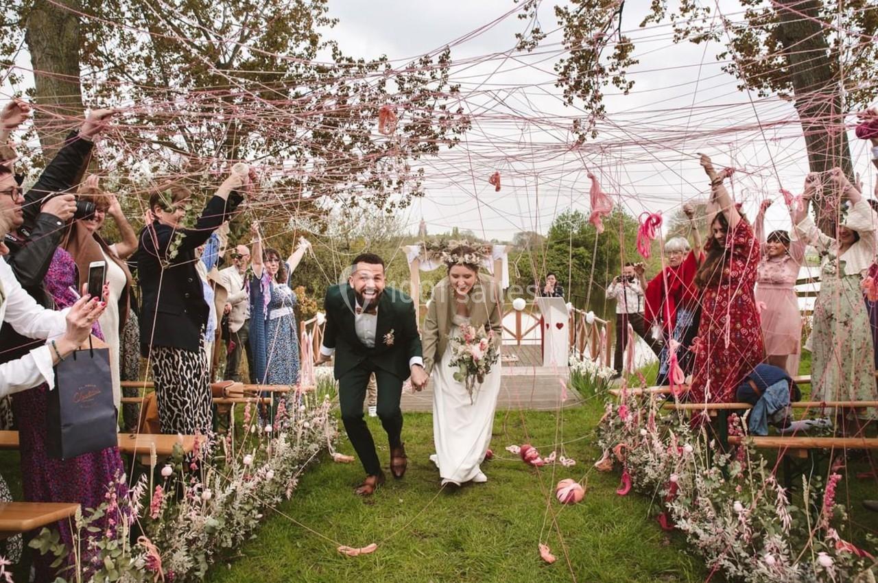 Un couple de mariés souriants traverse une allée décorée de fils colorés, entouré d'invités célébrant.