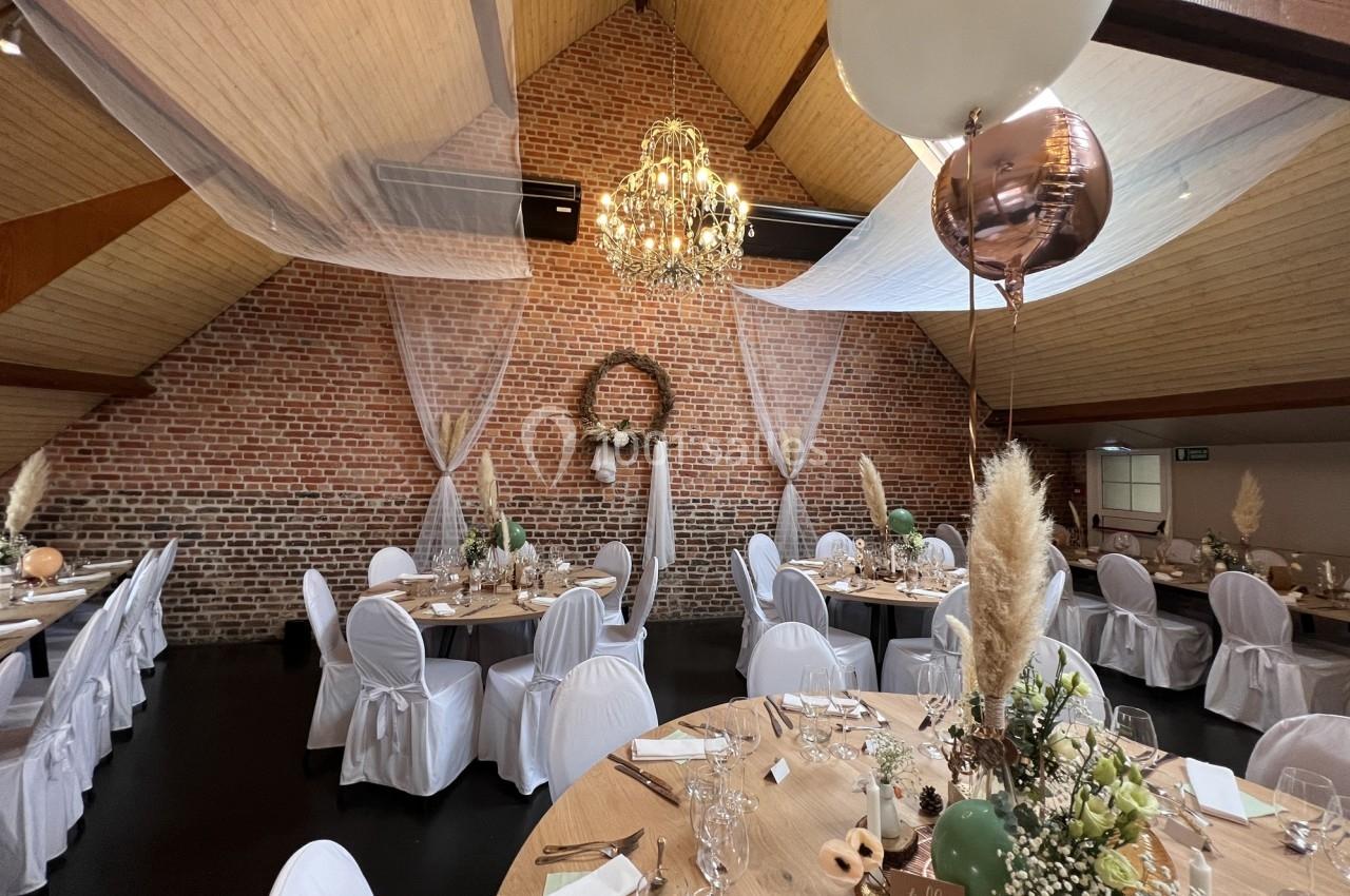 Salle de réception décorée avec des tables rondes, nappes blanches, ballons, fleurs et un lustre suspendu.
