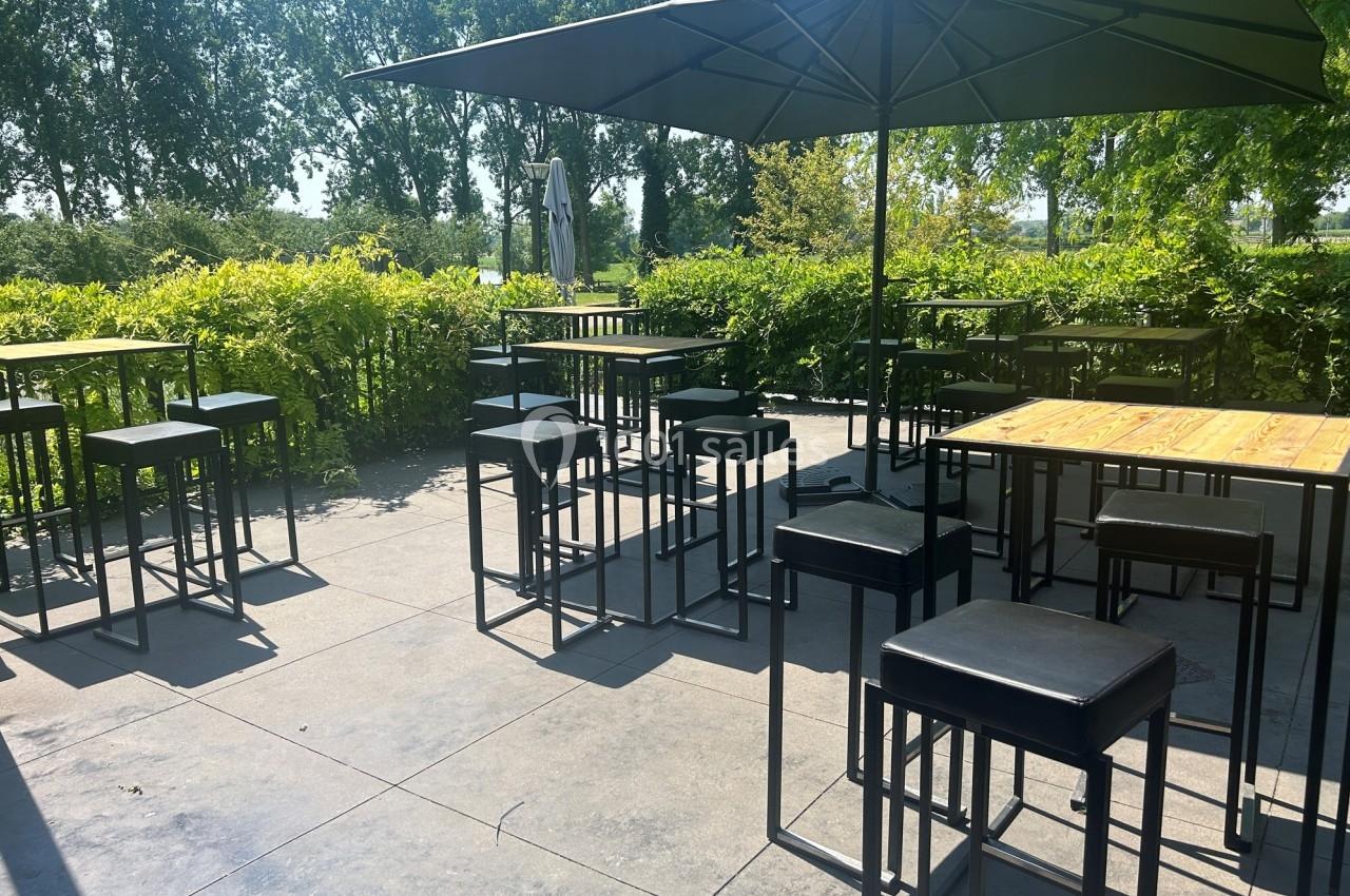Terrasse extérieure avec tables hautes, tabourets noirs et parasols, entourée de verdure et baignée de lumière naturelle.