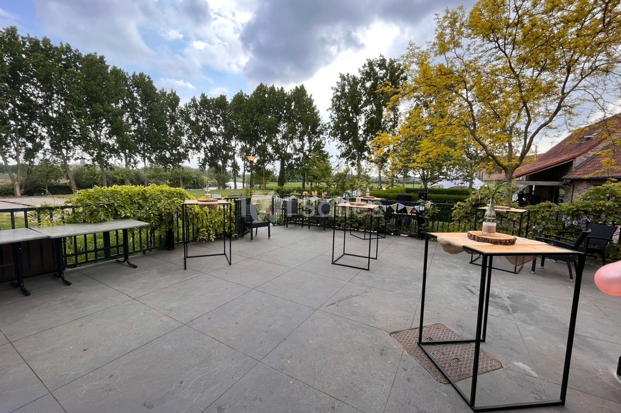 Terrasse extérieure avec tables espacées, entourée de verdure et d'arbres sous un ciel partiellement nuageux.