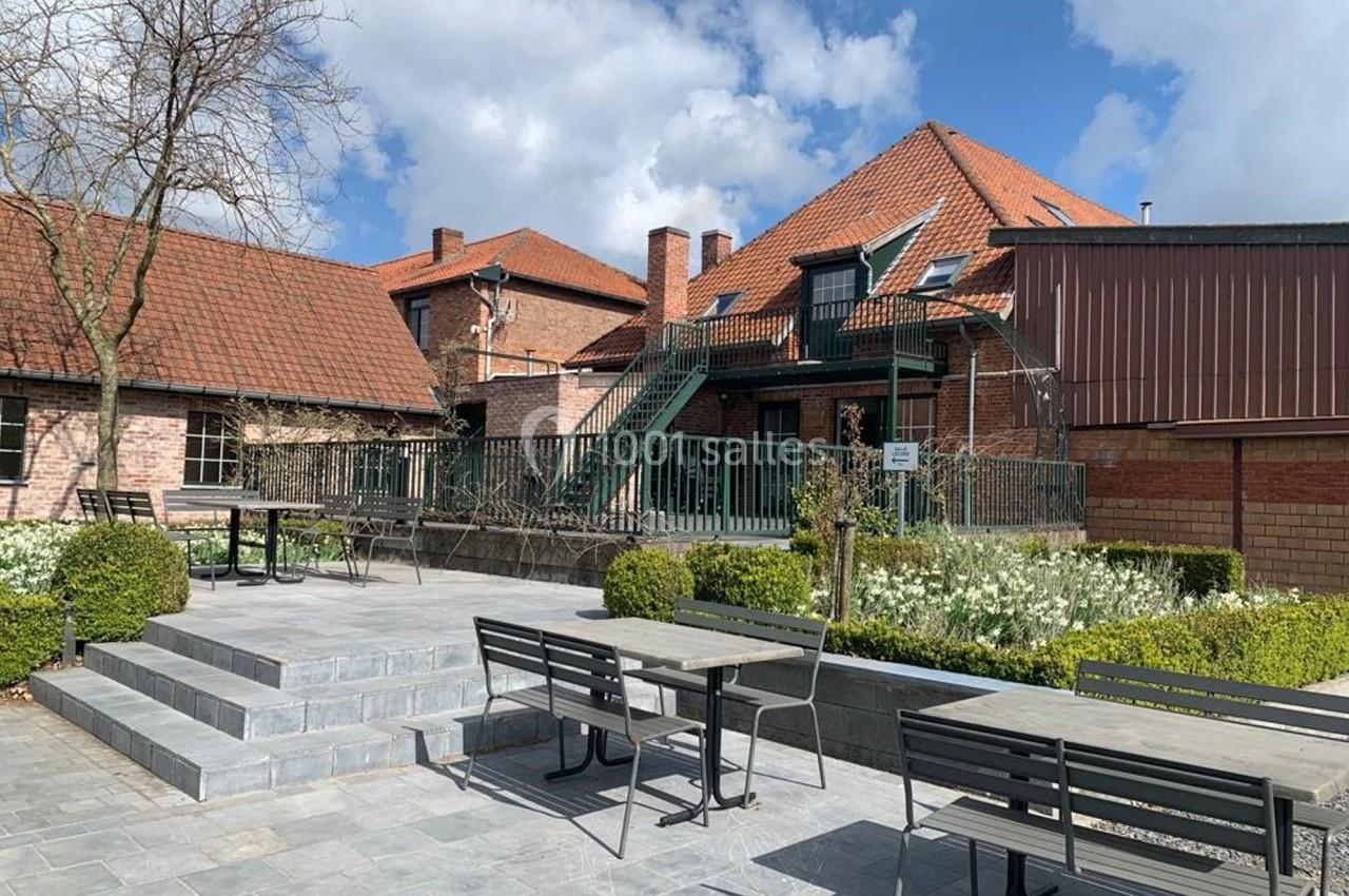 Cour extérieure pavée avec tables, chaises, escaliers en métal et bâtiments en briques rouges sous un ciel partiellement…