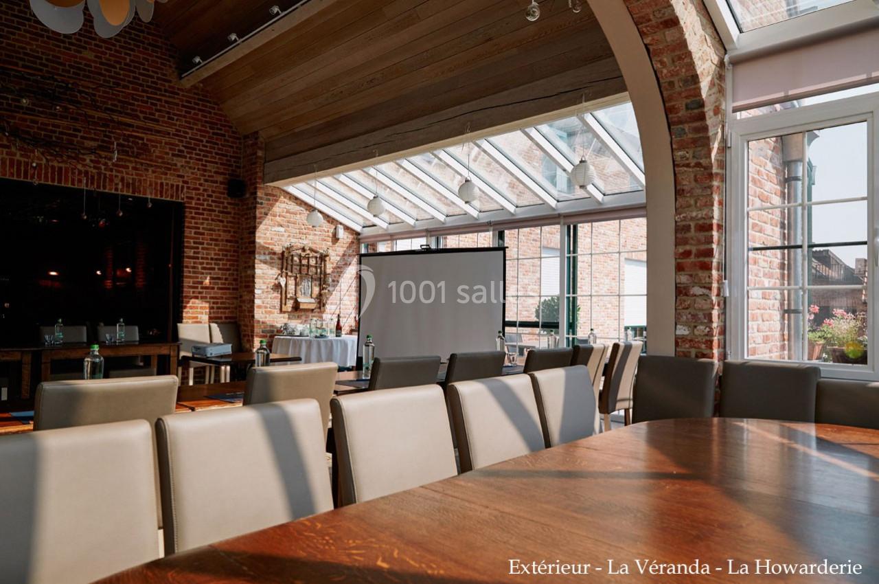 Salle lumineuse avec murs en briques, grandes baies vitrées, table en bois et chaises alignées devant un écran de projection.