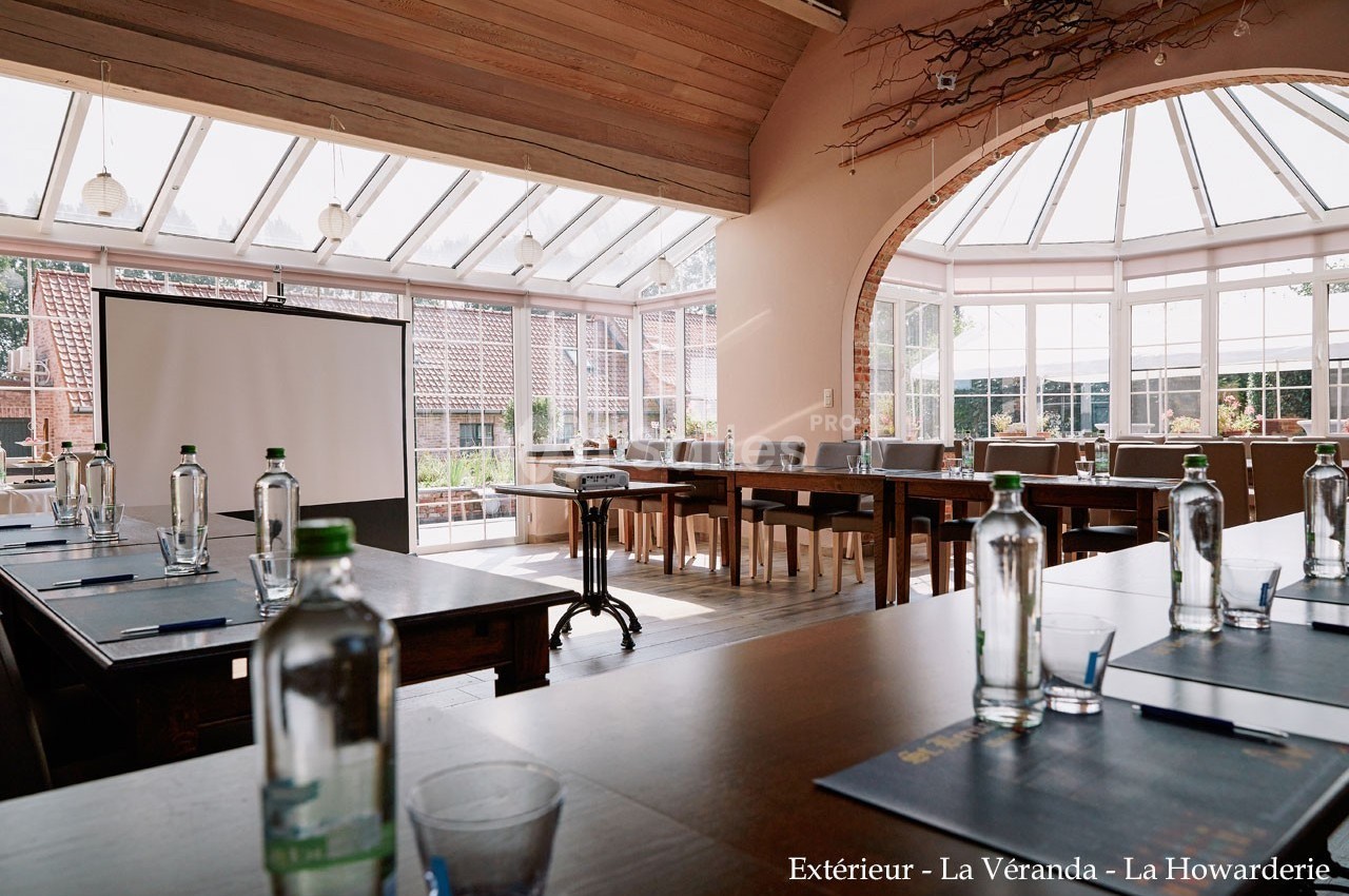 Salle de réunion lumineuse avec grandes baies vitrées, tables en bois, bouteilles d'eau et écran de projection.