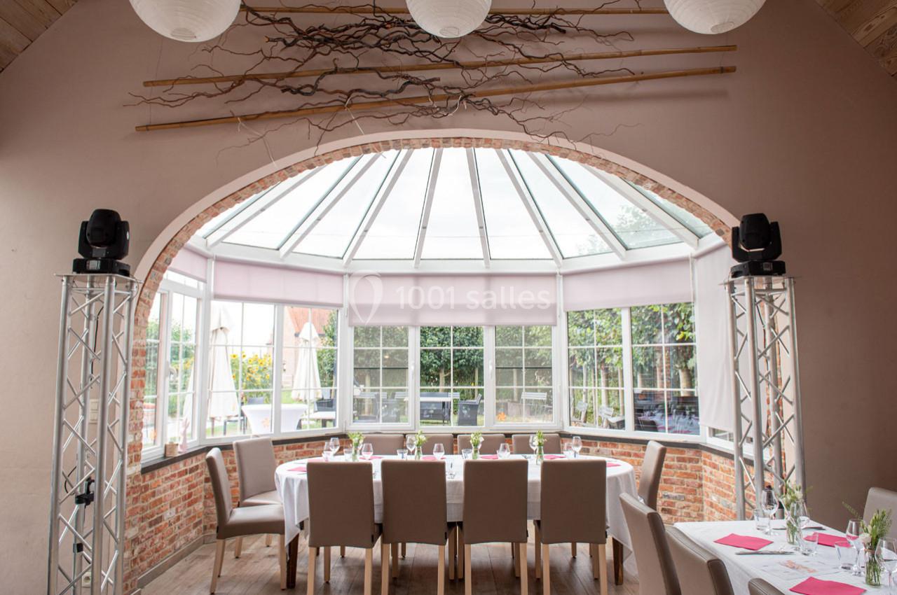 Salle lumineuse avec une grande verrière, des tables dressées, des chaises beige et une décoration naturelle en branches.