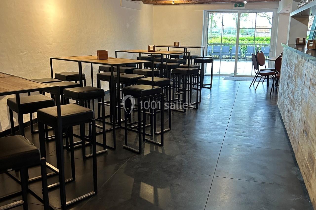 Salle lumineuse avec tables hautes, tabourets noirs, sol en béton et plafond en bois, donnant sur une baie vitrée.