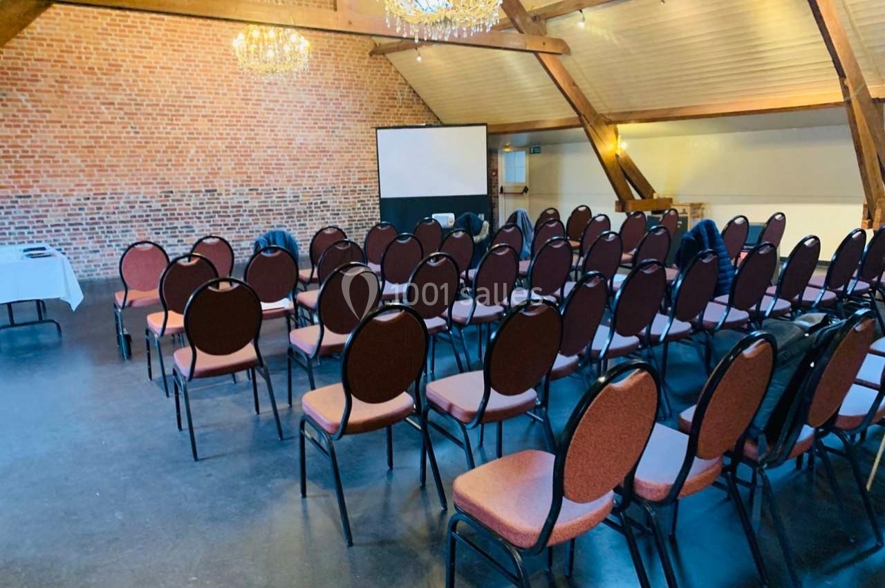 Salle de conférence avec des rangées de chaises vides, un écran de projection et des murs en briques apparentes.