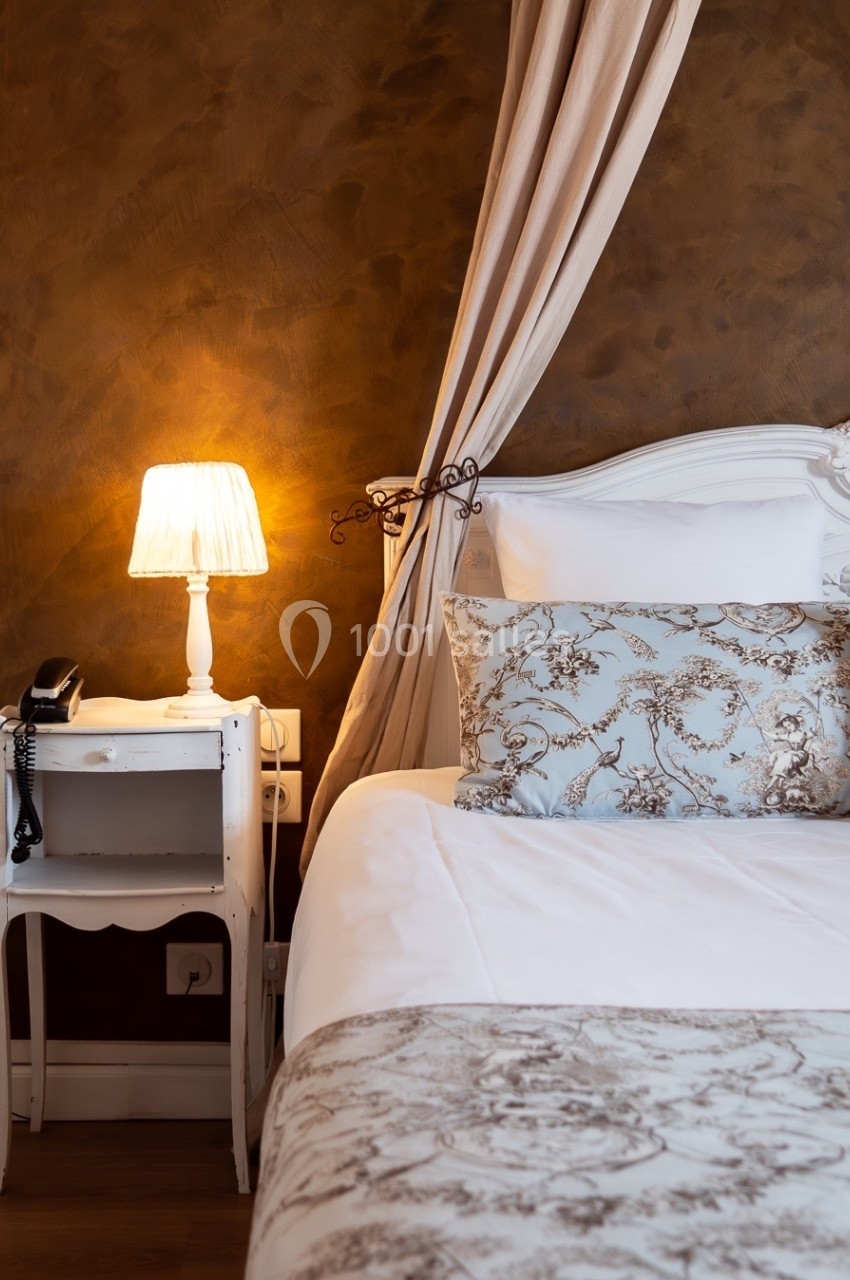 Lit avec tête de lit blanche, draps à motifs floraux, table de chevet avec lampe allumée et téléphone noir.