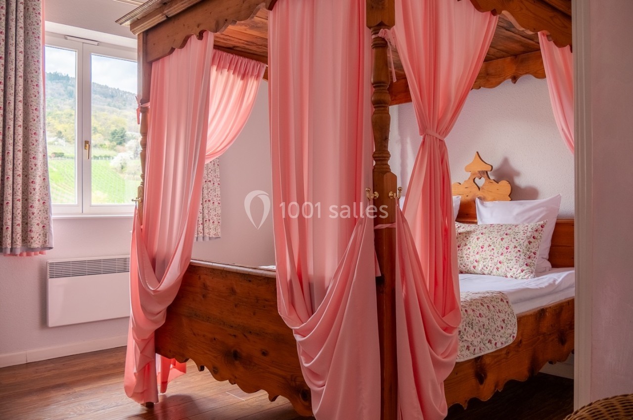 Lit à baldaquin en bois avec rideaux roses dans une chambre lumineuse, vue sur un paysage extérieur par la fenêtre.