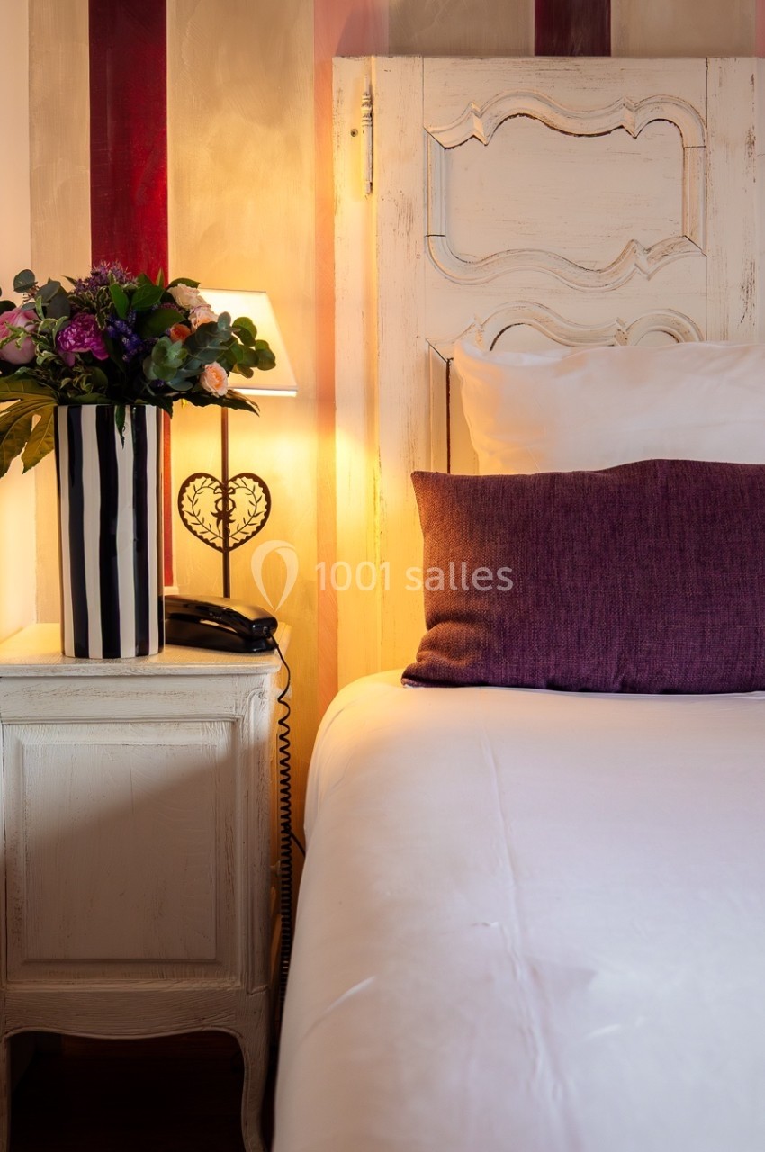 Table de chevet avec lampe allumée, vase de fleurs et téléphone, à côté d'un lit avec oreiller violet.