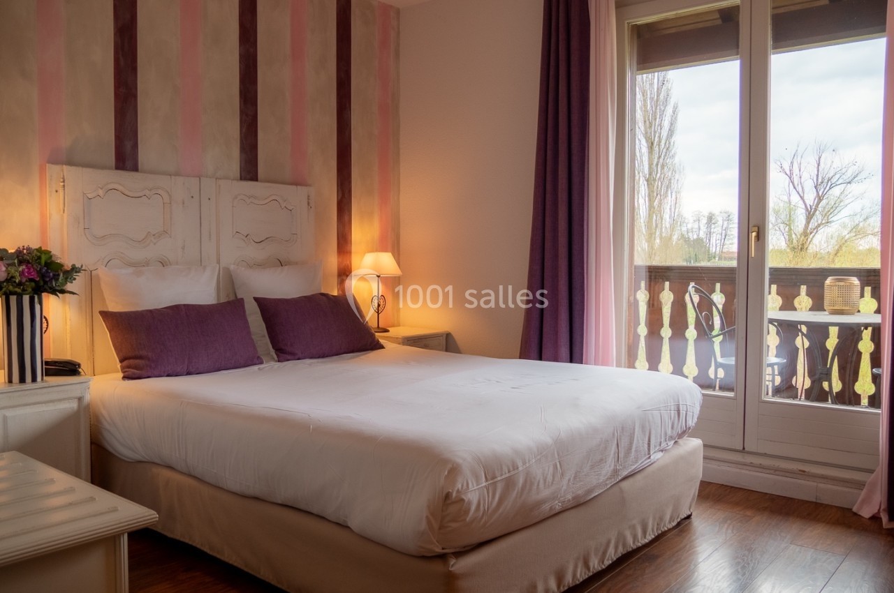 Chambre lumineuse avec lit double, murs à rayures roses, lampes de chevet et balcon donnant sur un paysage arboré.