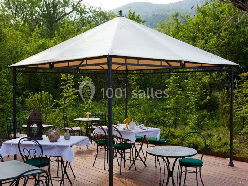 Tonnelle blanche sur une terrasse en bois avec tables et chaises, entourée de verdure et d'arbres.