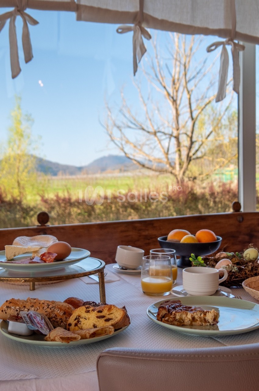 Petit-déjeuner servi sur une table près d'une fenêtre avec vue sur un paysage champêtre ensoleillé.