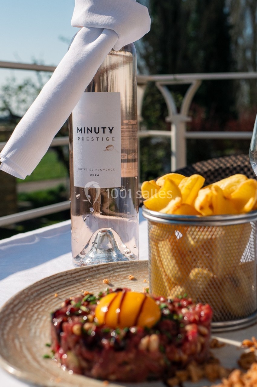 Bouteille de vin rosé, tartare de viande avec œuf, frites et verre sur une table en extérieur ensoleillé.