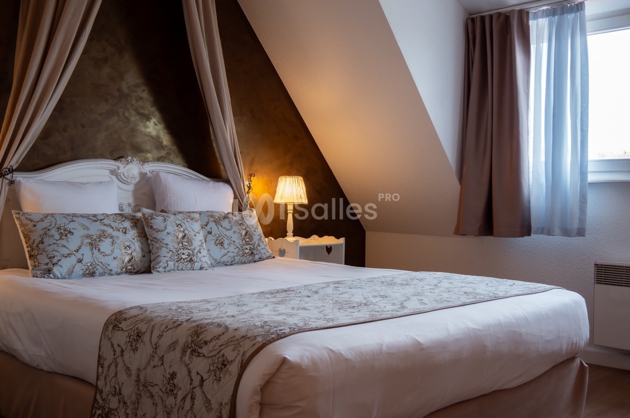 Lit double avec tête de lit sculptée, draps blancs et coussins décoratifs, dans une chambre mansardée éclairée par une lampe.