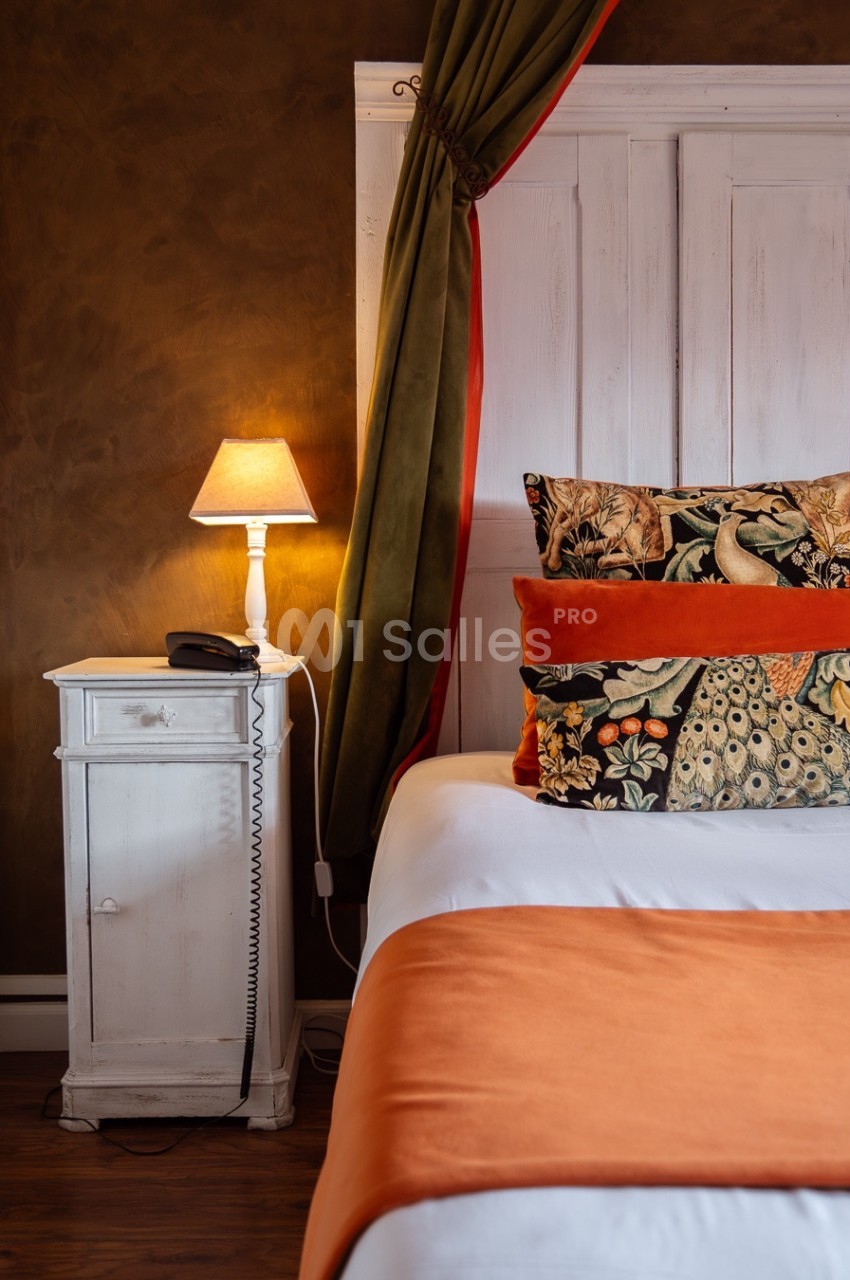 Lit avec coussins décoratifs colorés, lampe allumée sur une table de chevet blanche dans une chambre chaleureuse.