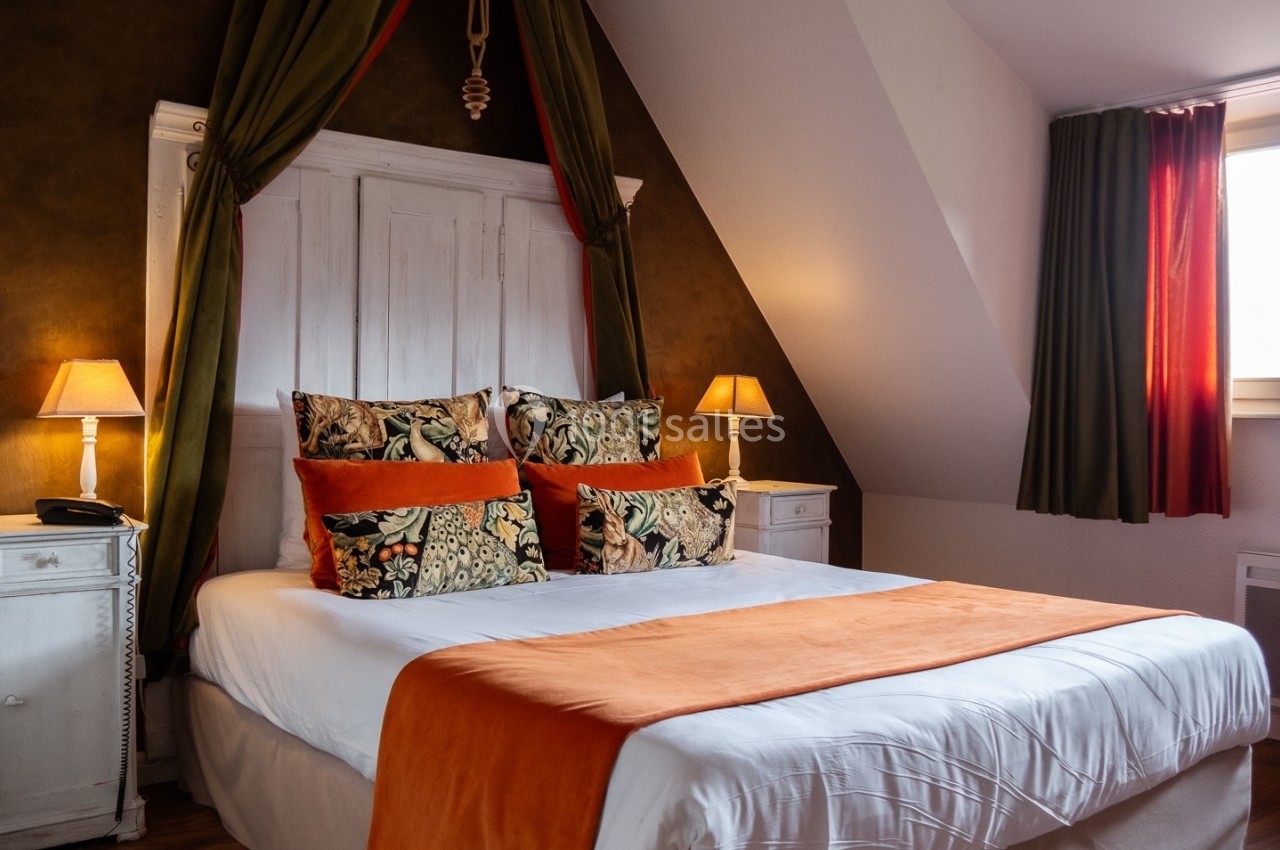 Lit double avec draps blancs, couverture orange, coussins décoratifs, encadré par deux lampes sur des tables de chevet…
