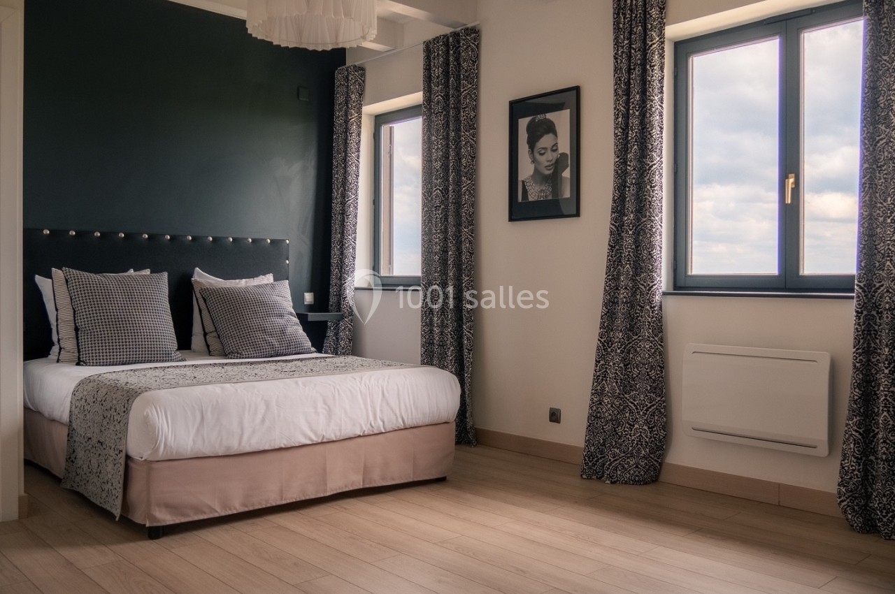 Chambre lumineuse avec un lit double, des rideaux à motifs, un mur sombre et deux fenêtres donnant sur l'extérieur.