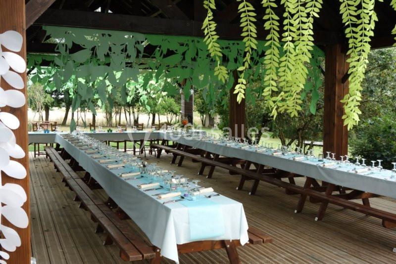 Tables en bois alignées et décorées sous une pergola ornée de guirlandes vertes, dans un cadre extérieur.