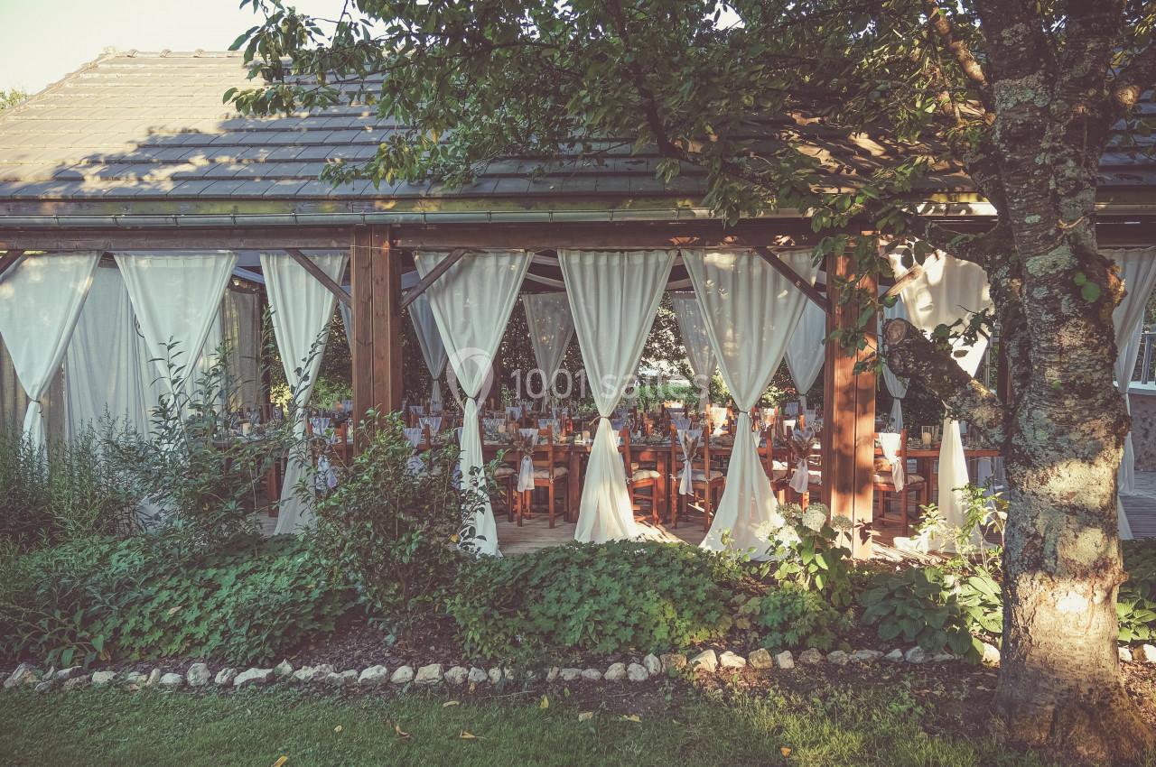 Salle de réception lumineuse avec des tables dressées, vue à travers des rideaux blancs et entourée de verdure.