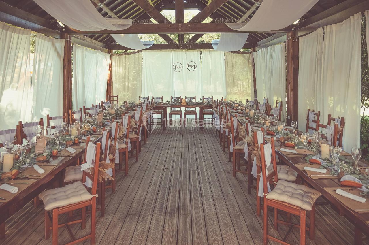 Salle de réception en bois décorée pour un repas, avec longues tables dressées et chaises alignées sous un toit ouvert.