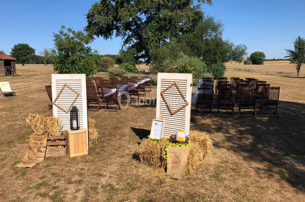 Décor champêtre avec bottes de paille, volets blancs, chaises en bois et arbres en arrière-plan sous un ciel bleu.