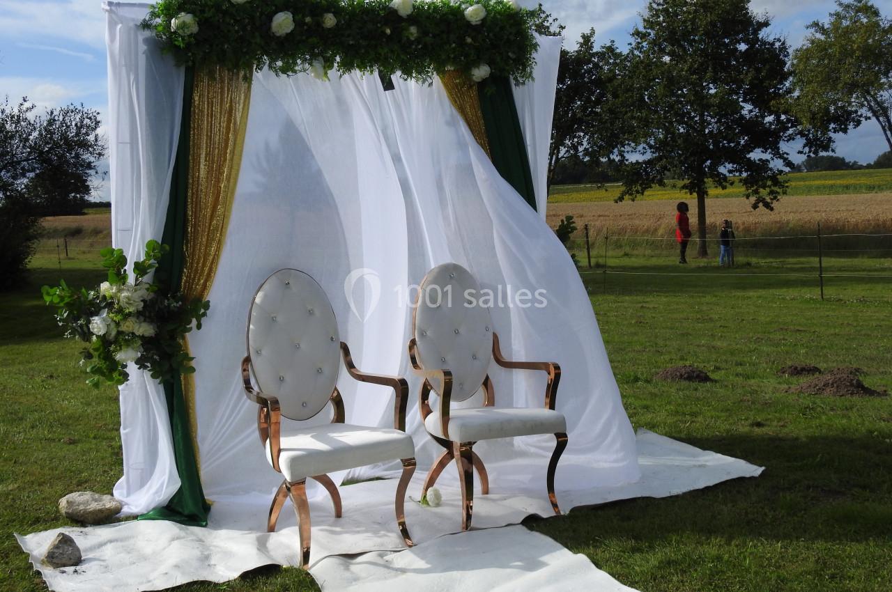 Deux fauteuils blancs devant une arche décorée de voilages, fleurs et feuillage, installée sur une pelouse.