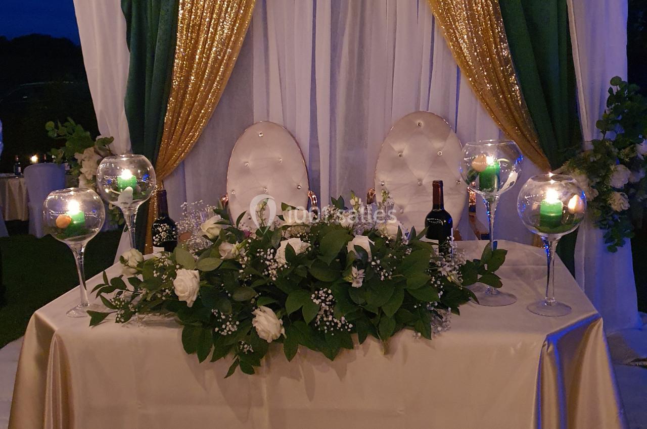 Table décorée pour un mariage avec nappes blanches, chandeliers, fleurs blanches et arche ornée de rideaux dorés et verts.