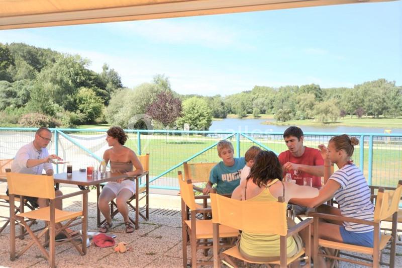 Des personnes assises à des tables en terrasse, discutant et partageant un moment convivial près d'un lac entouré de verdure.