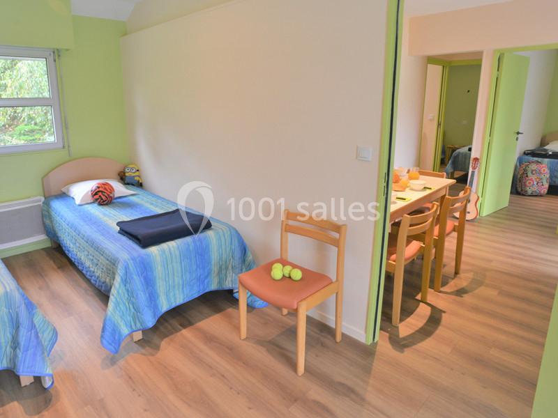 Chambre avec deux lits simples, parquet, murs verts, et une table à manger visible dans une pièce adjacente.