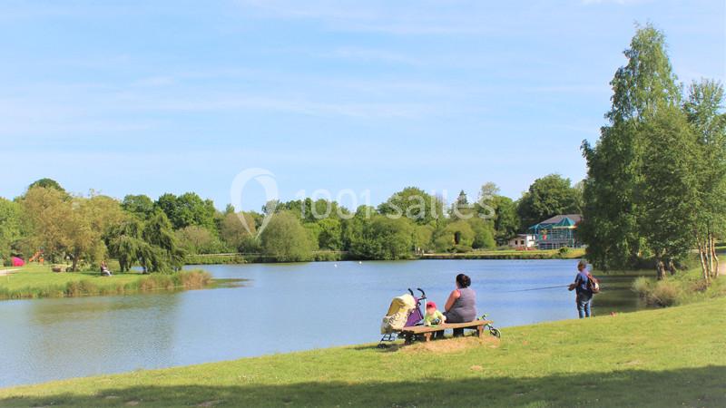 Deux personnes assises près d'un lac entouré d'arbres, avec une poussette et une personne pêchant à proximité.