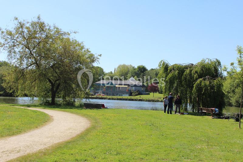 Un parc verdoyant avec un sentier, un lac, des arbres et quelques personnes près de l'eau sous un ciel dégagé.