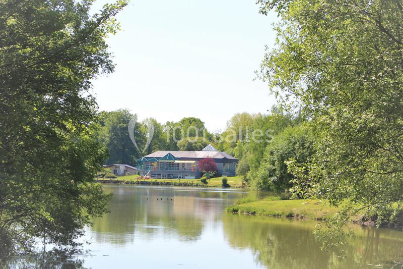 Vue d'un bâtiment entouré de verdure au bord d'un étang calme, sous un ciel dégagé.