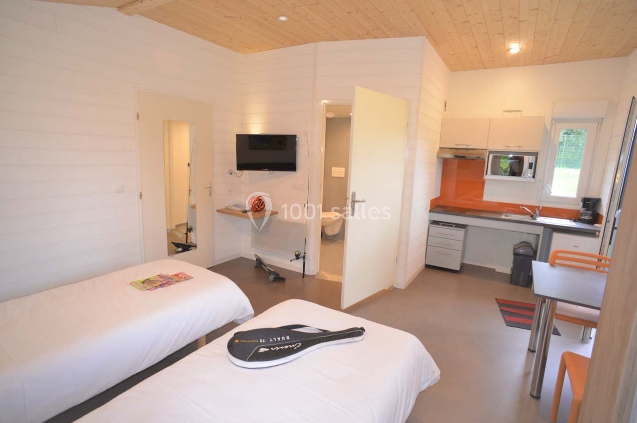 Intérieur d'un chalet avec deux lits simples, une kitchenette équipée, une table et une télévision fixée au mur.