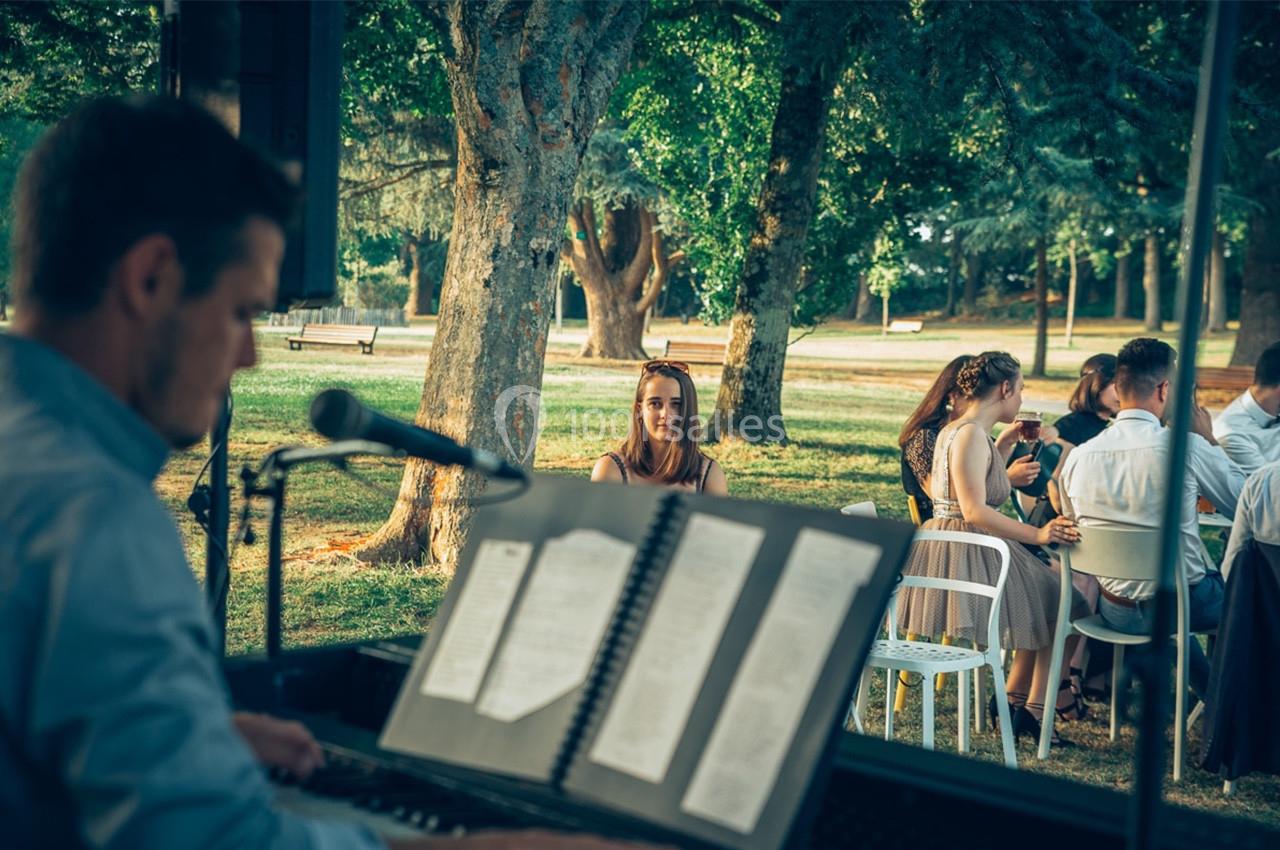 Un musicien joue du piano en plein air tandis que des personnes sont assises sous des arbres dans un parc.