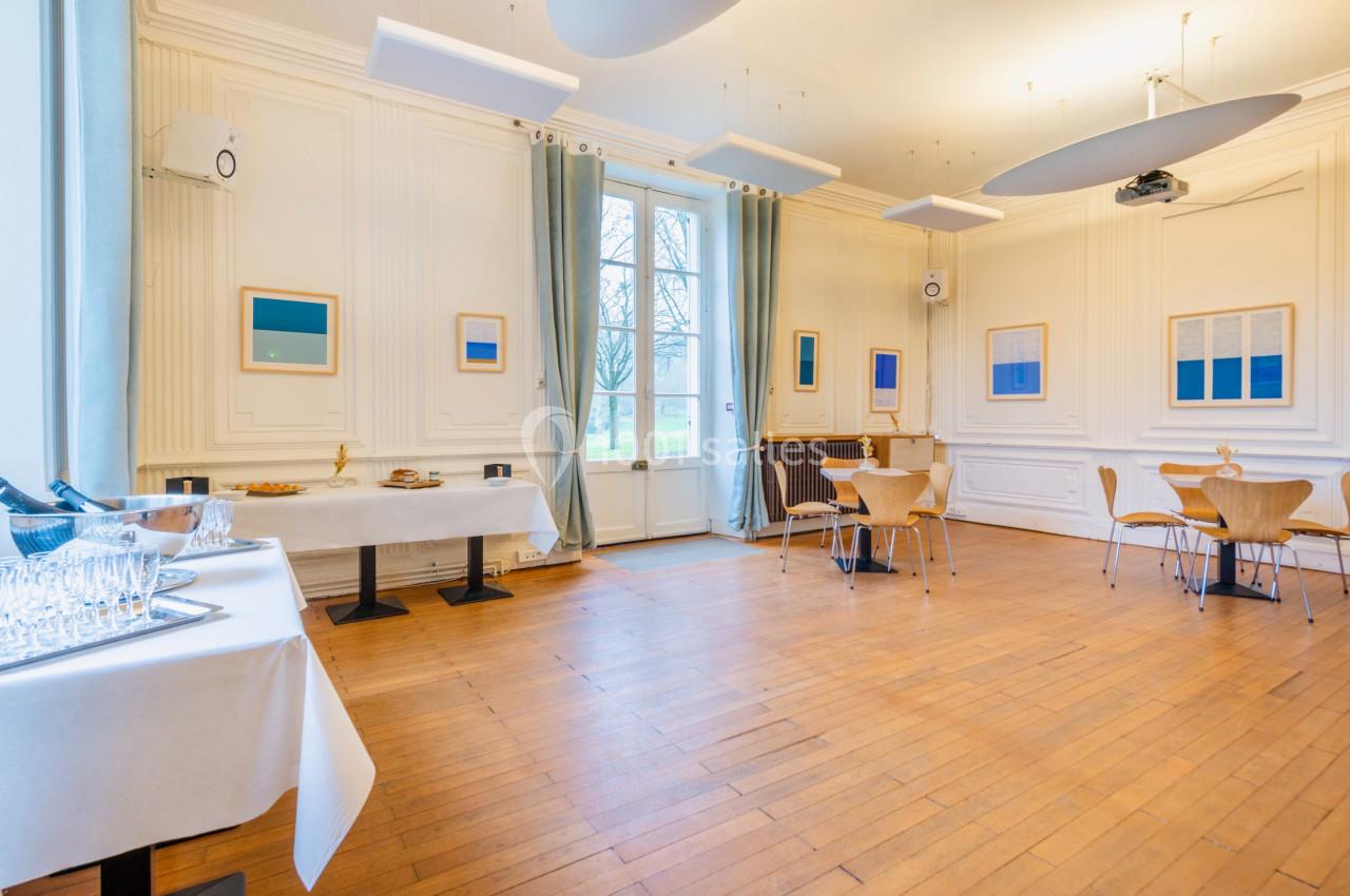 Salle lumineuse avec parquet, tables et chaises en bois, décorée de tableaux modernes et grandes fenêtres donnant sur un…