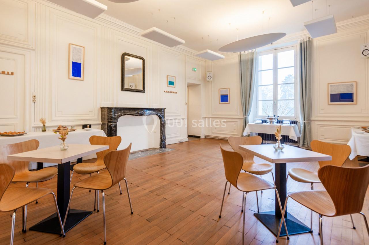 Salle lumineuse avec parquet en bois, tables et chaises modernes, cheminée et grandes fenêtres donnant sur l'extérieur.