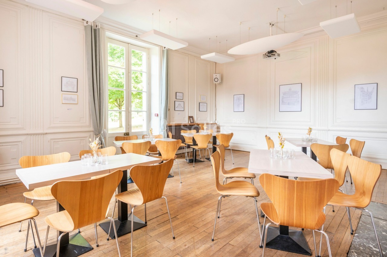 Salle lumineuse avec parquet, tables blanches, chaises en bois clair et grandes fenêtres donnant sur un extérieur verdoyant.