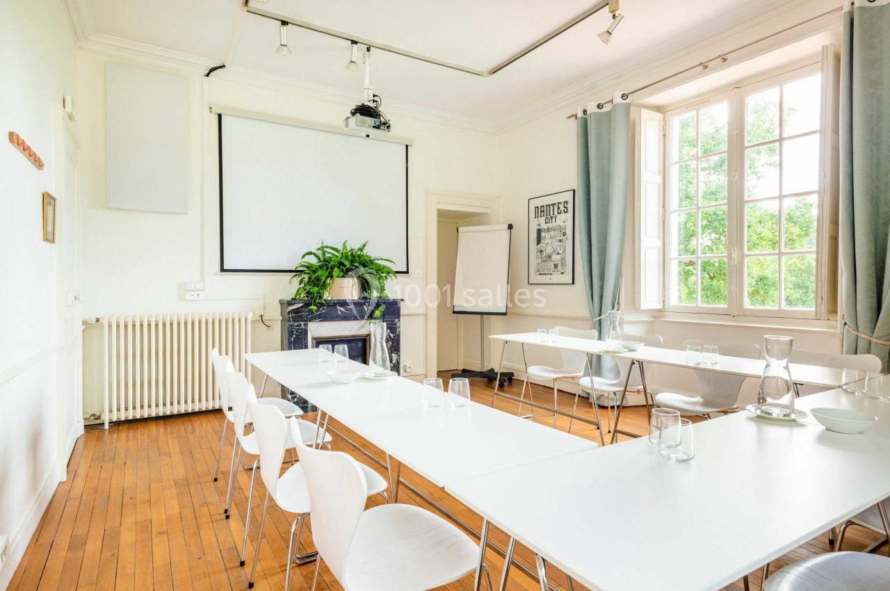 Salle lumineuse avec tables blanches alignées, chaises, tableau blanc, projecteur, plantes et grande fenêtre donnant sur un…