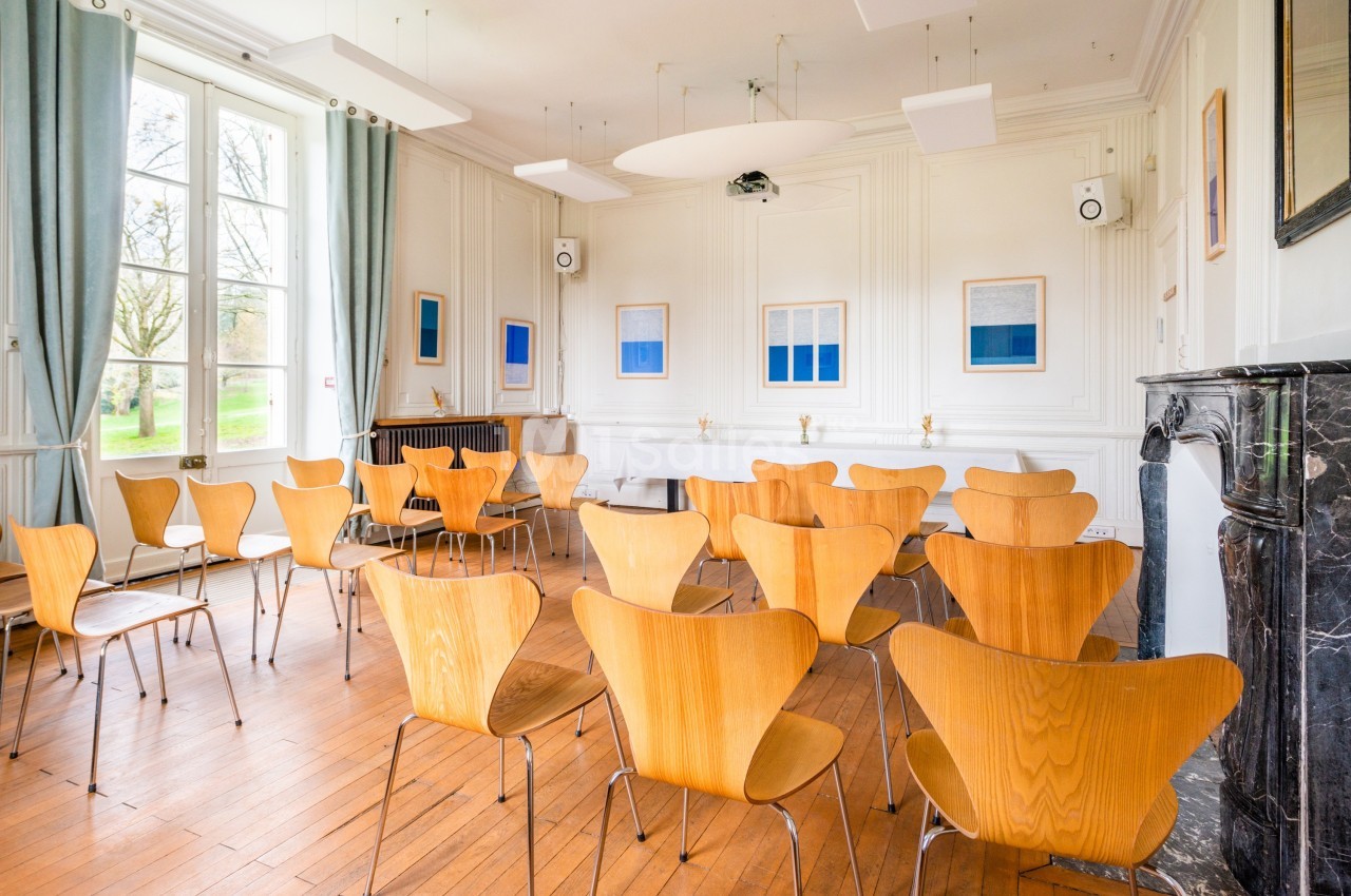 Salle lumineuse avec des chaises en bois disposées face à un écran, parquet au sol et grandes fenêtres donnant sur un jardin.