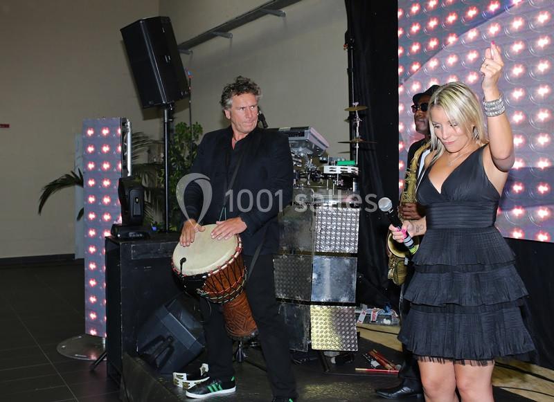 Un homme joue du djembé et une femme chante sur scène, entourés d'équipements lumineux et d'un saxophoniste.