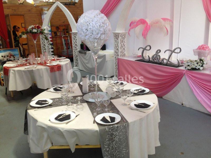 Salle décorée pour un événement avec tables dressées, nappes blanches, chemins de table gris et roses, et ornements floraux.