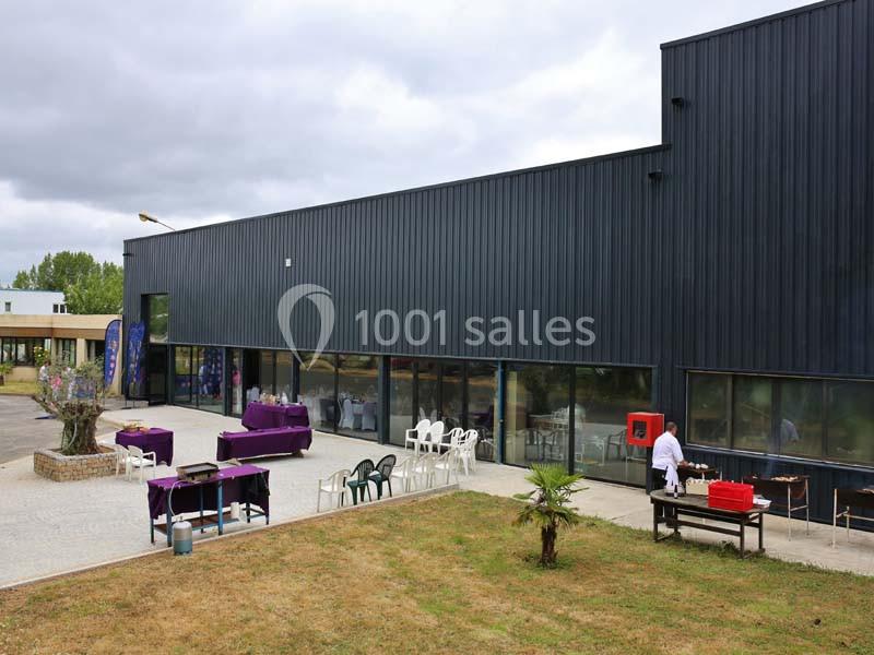 Vue d'un bâtiment moderne avec façade sombre, tables dressées en extérieur et un homme près d'un barbecue.