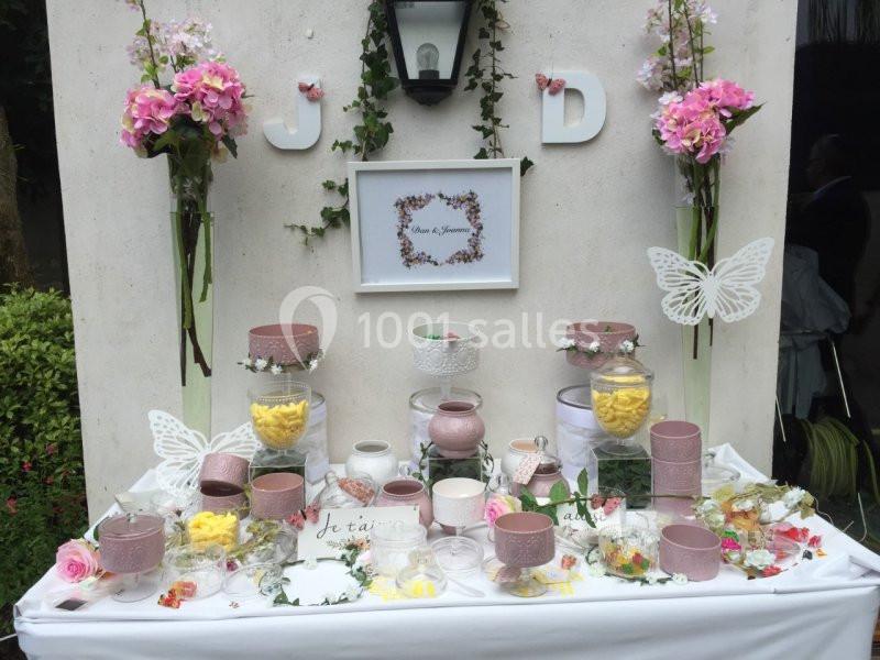 Table décorée pour un événement avec bonbonnières, fleurs roses, papillons et initiales ’J’ et ’D’ accrochées au mur.