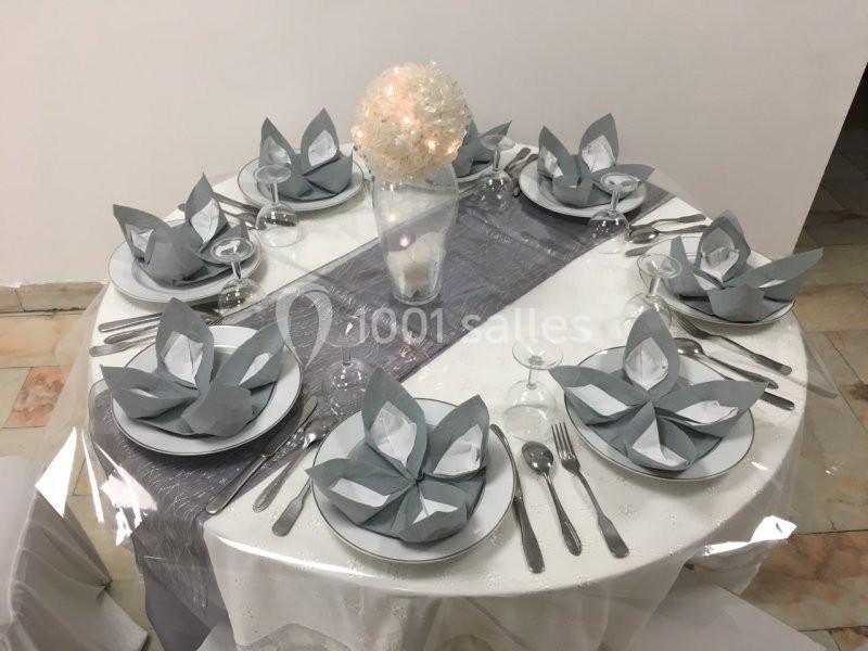 Table ronde élégamment dressée avec des serviettes pliées en forme de fleurs, un chemin de table gris et un centre de table…