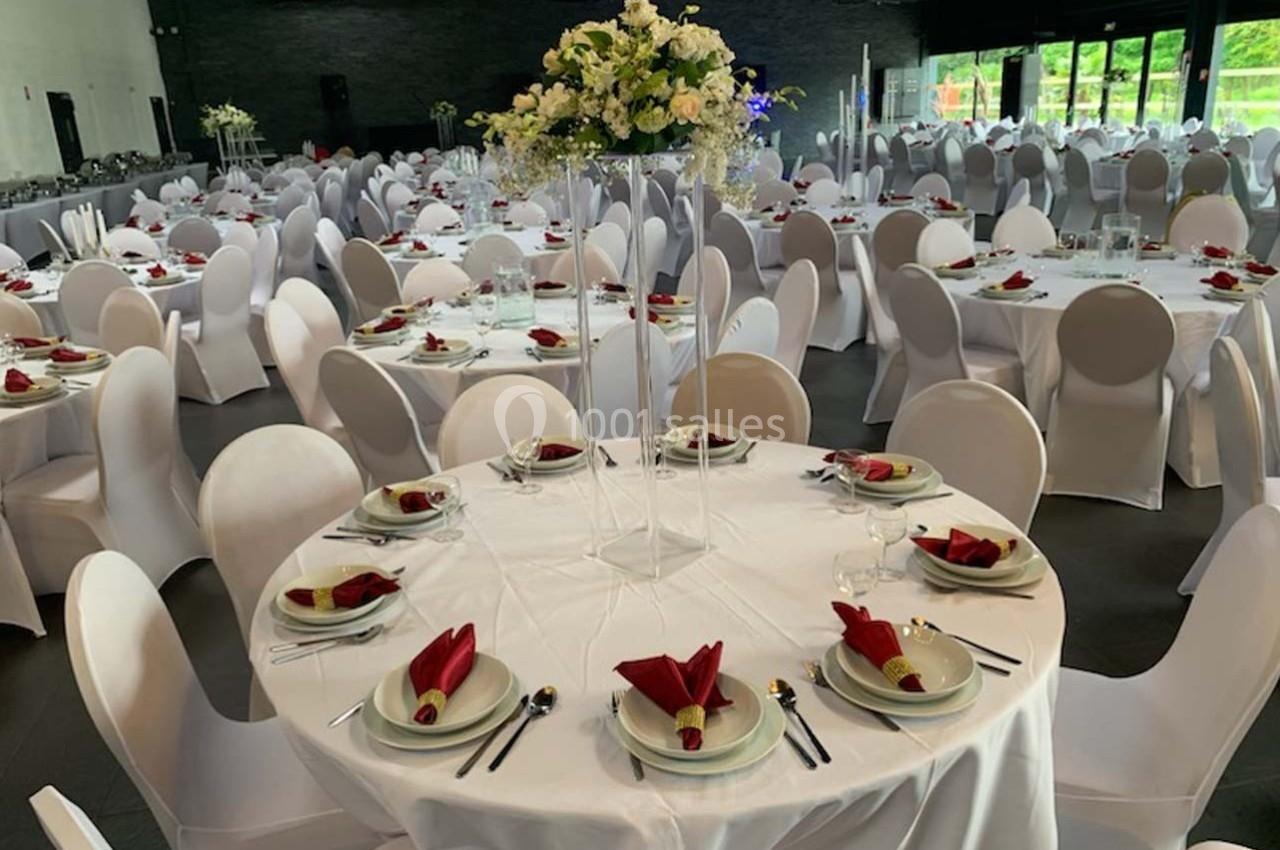 Salle de réception décorée avec des tables rondes, nappes blanches, serviettes rouges pliées et centres de table floraux.