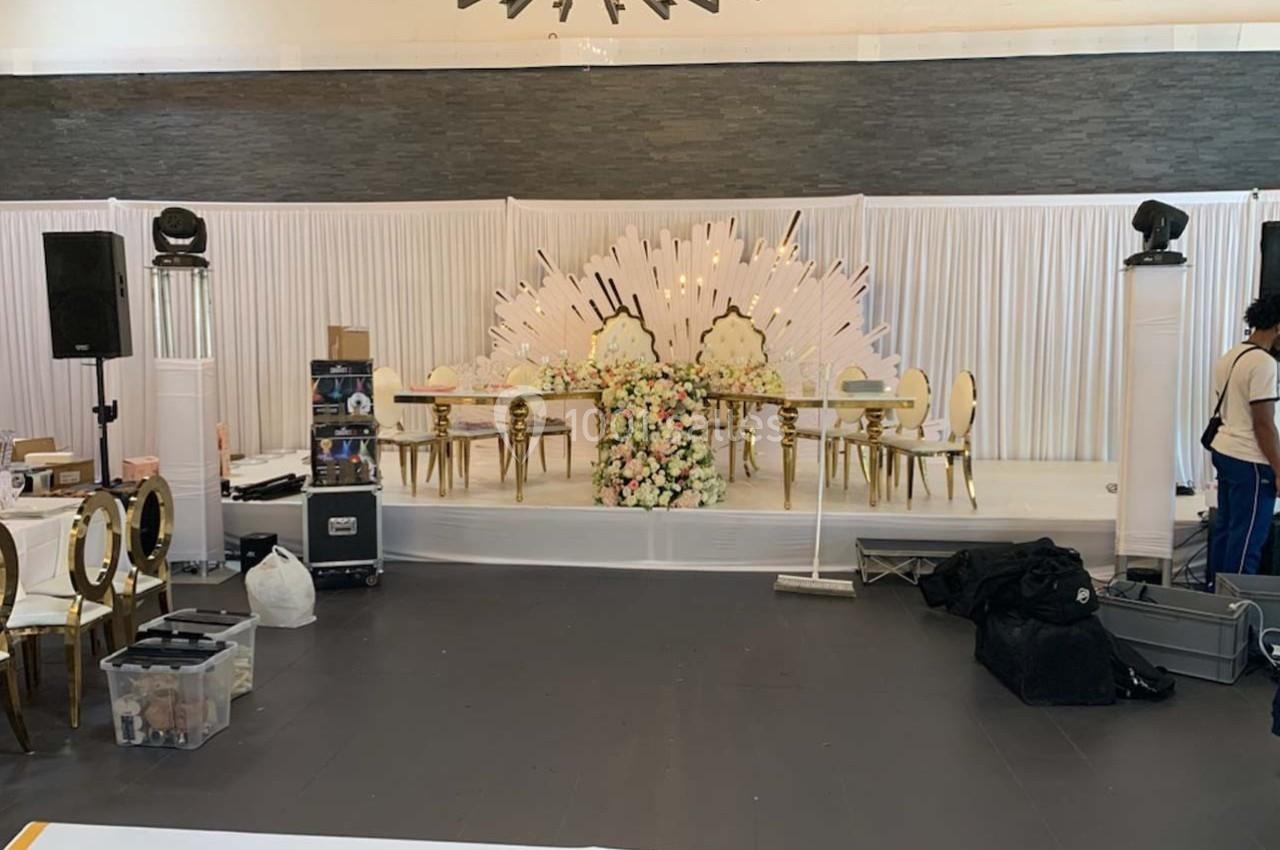 Scène décorée pour un événement avec une table ornée de fleurs, des chaises dorées et un fond blanc lumineux.