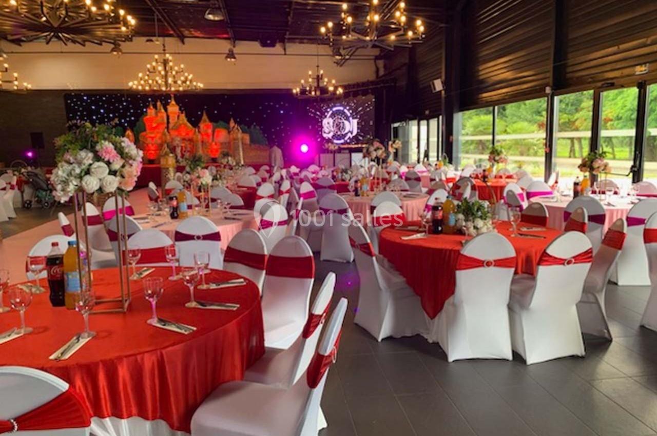 Salle de réception décorée avec des tables rondes nappées de rouge, chaises blanches et éclairage chaleureux.