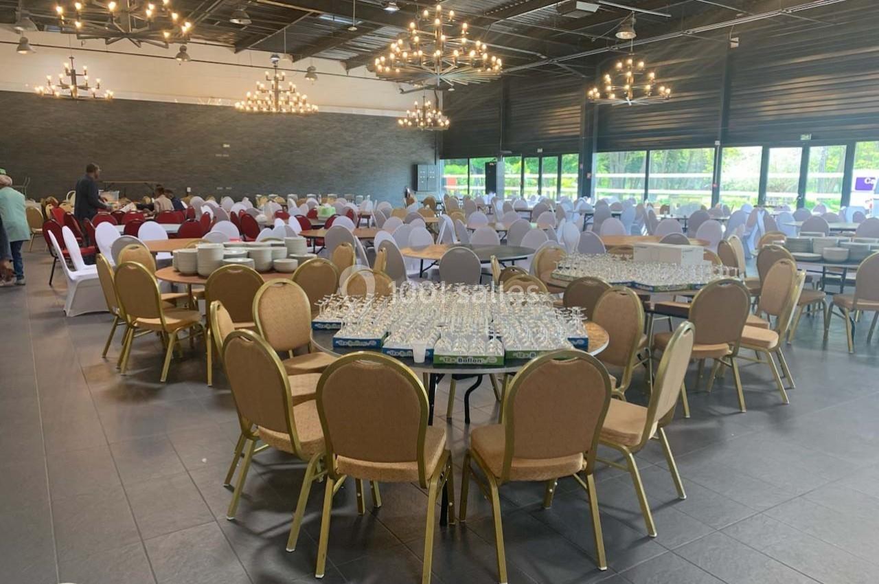 Salle de réception avec tables rondes dressées de verres et chaises disposées, éclairée par des lustres suspendus.