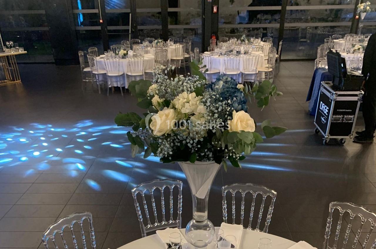 Centre de table floral avec roses blanches et feuillage, dans une salle de réception élégante avec tables dressées.