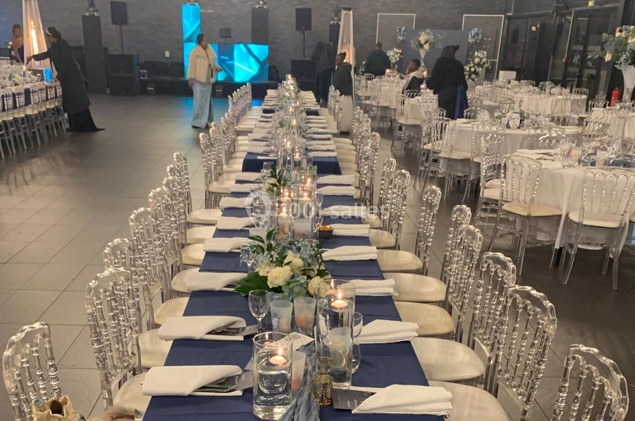 Tables décorées pour un événement, alignées avec nappes bleues, bougies, fleurs et chaises transparentes dans une salle.